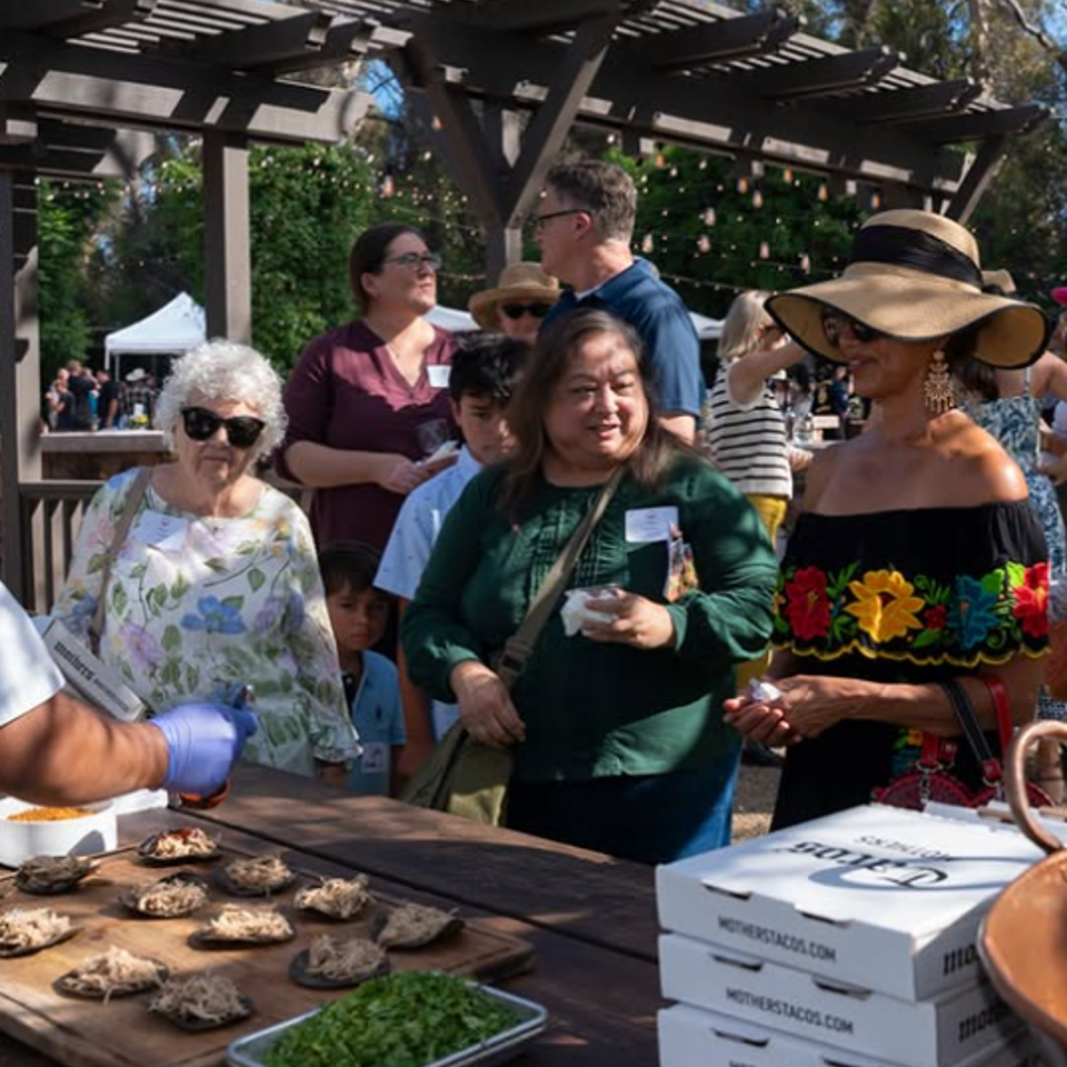 Visitors gather at an outdoor food stall beneath a pergola, while grilled food, herbs, and stacked takeaway boxes fill the table.