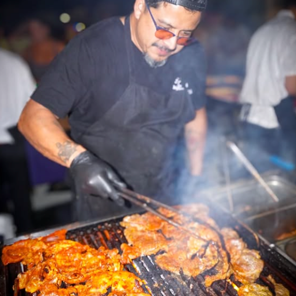 Grill cook wearing gloves and a cap tends sizzling marinated meats over a smoky barbecue grill at night.