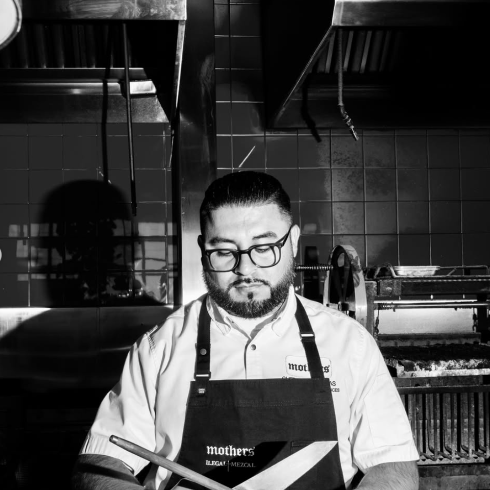 Bearded chef in glasses and gloves sharpens a knife in a tiled restaurant kitchen beside the grill.