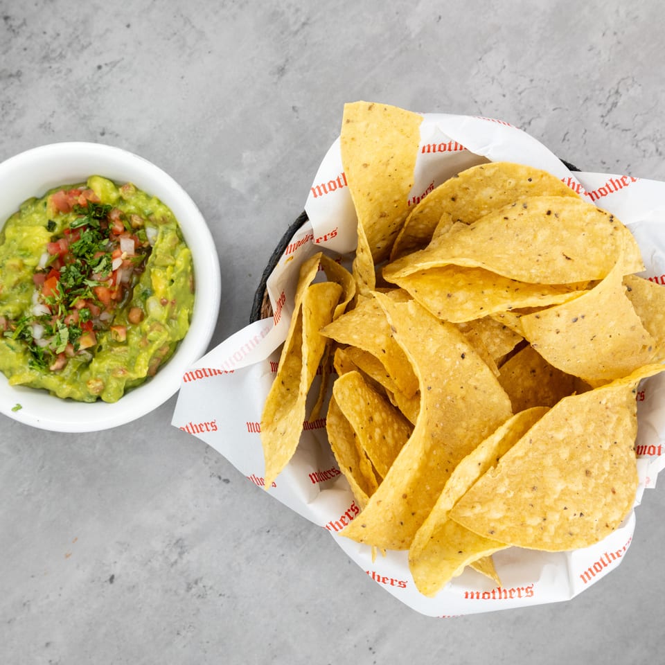Basket of golden tortilla chips beside a bowl of chunky guacamole topped with tomatoes, onions, and cilantro.