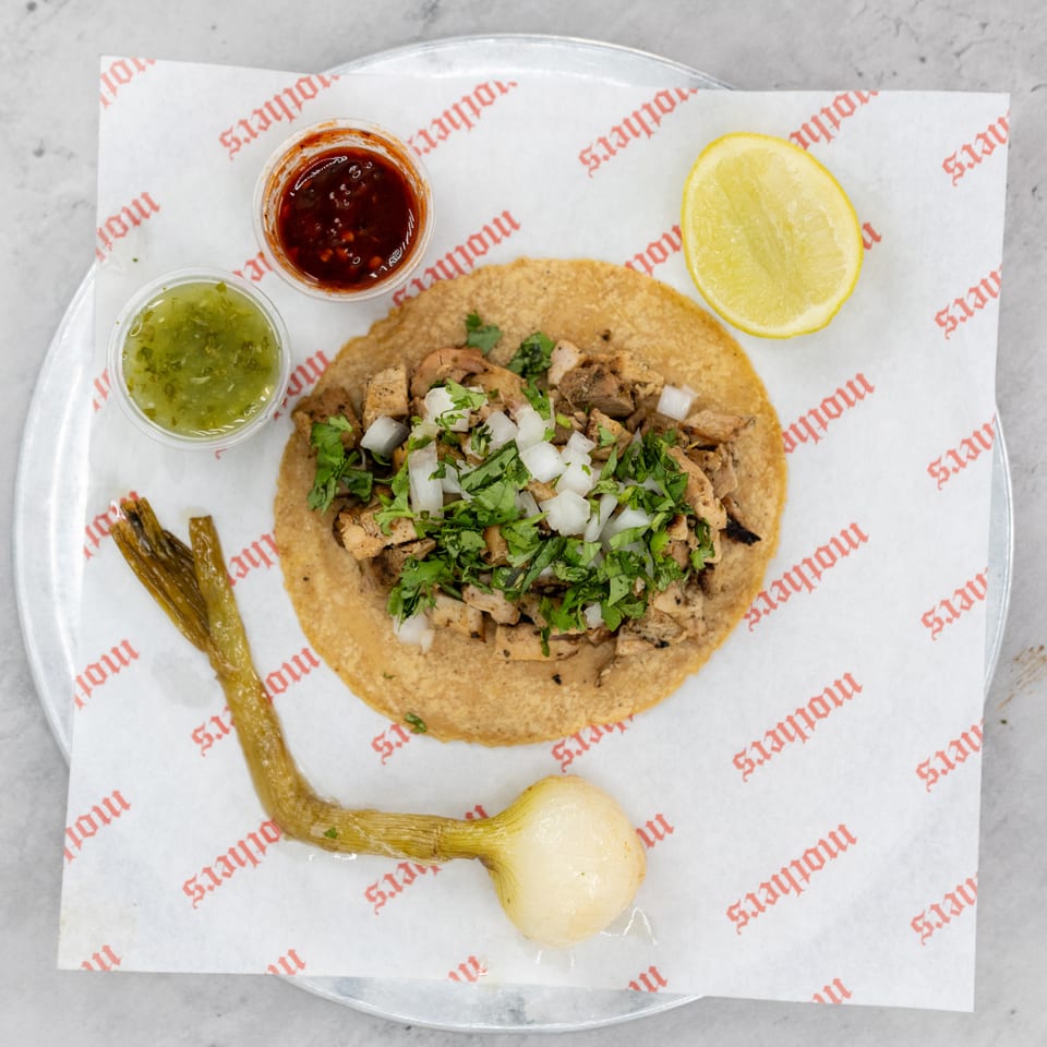 Overhead view of a meat taco topped with cilantro and onion, served with lime, grilled peppers, onion, and two sauces.