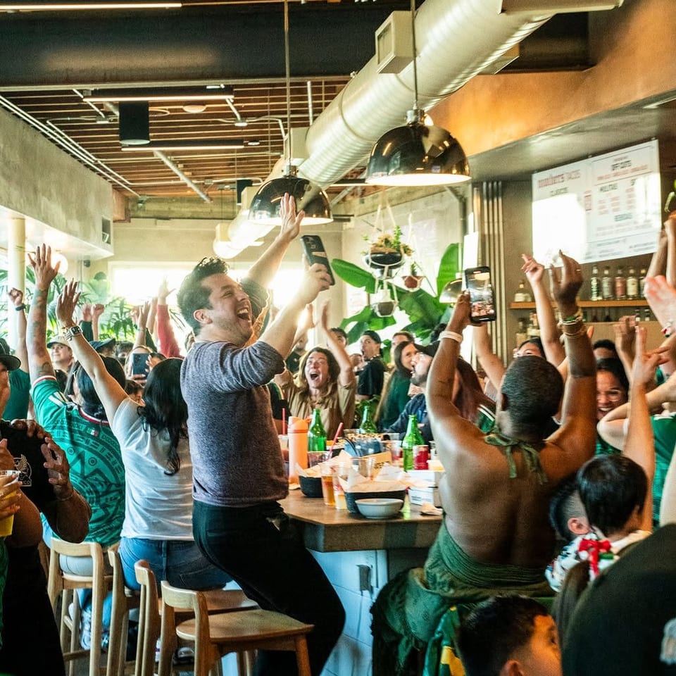 Crowded restaurant patrons cheer and raise their phones around a dining table beneath industrial pendant lights.
