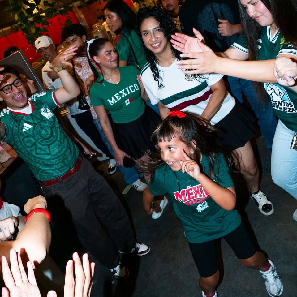 Diners in Mexico soccer jerseys celebrate and dance together inside a lively, warmly lit restaurant.