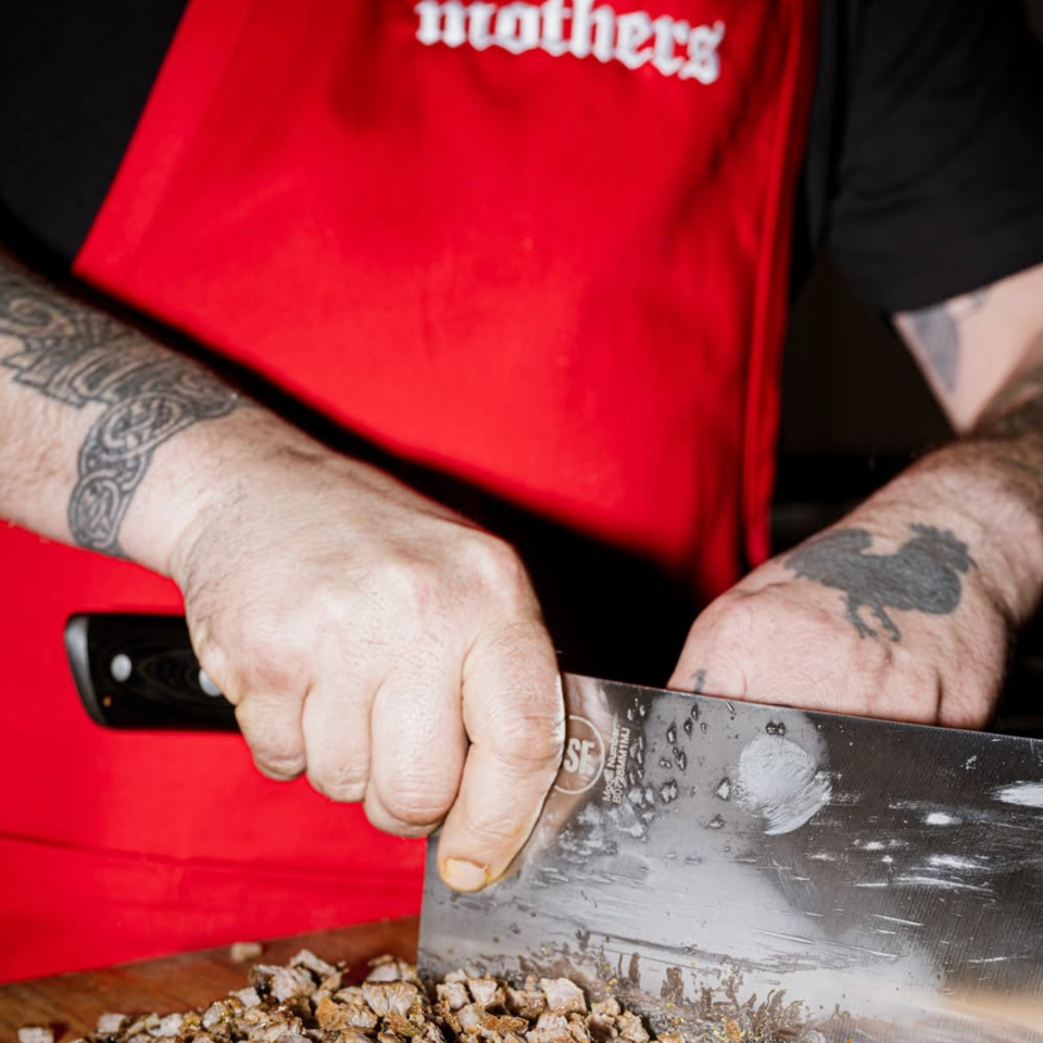 Tattooed chef in a red “Meathers” apron chops cooked meat with a large knife on a wooden cutting board.