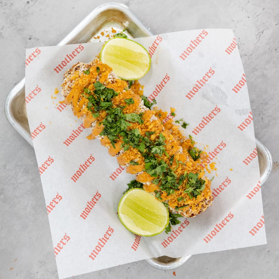 Crispy tacos topped with orange sauce, cotija cheese, cilantro, and lime wedges on branded restaurant paper.