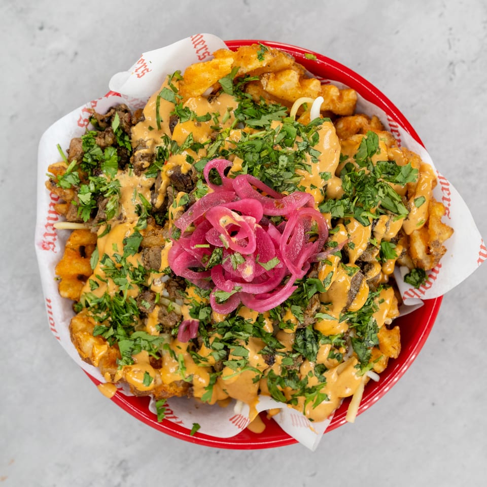 Loaded red basket of fries topped with grilled meat, orange sauce, chopped herbs, melted cheese, and pickled onions.