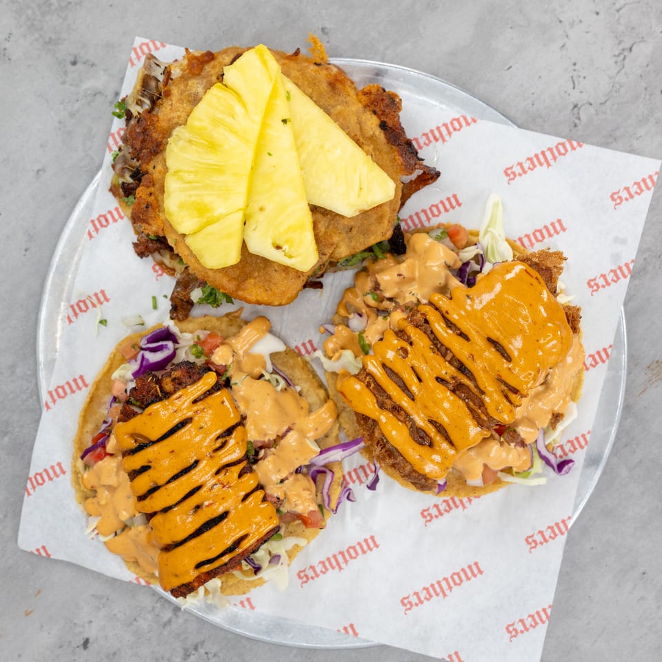 Overhead view of three loaded tacos with crispy fillings, shredded cabbage, creamy orange sauce, and pineapple slices on a branded tray.