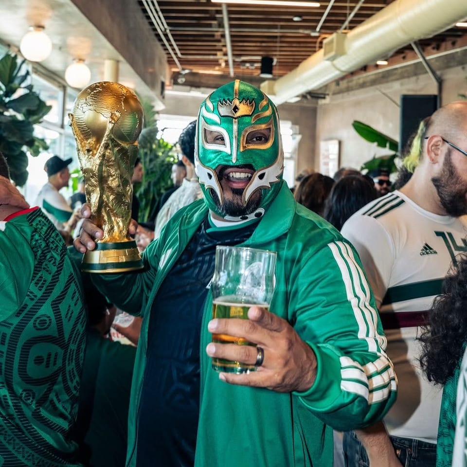 Crowded restaurant celebration featuring a fan in a green wrestling mask holding a trophy and glass of beer.