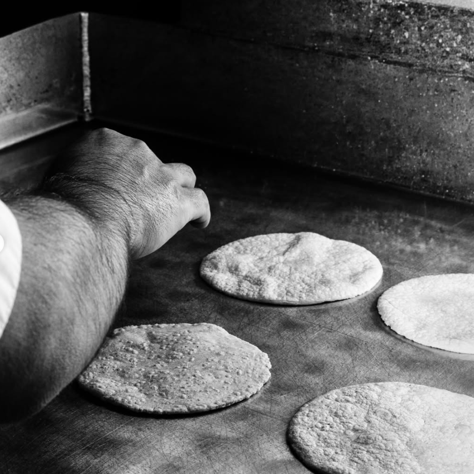 A cook’s hand prepares several round corn tortillas on a well-used metal griddle.