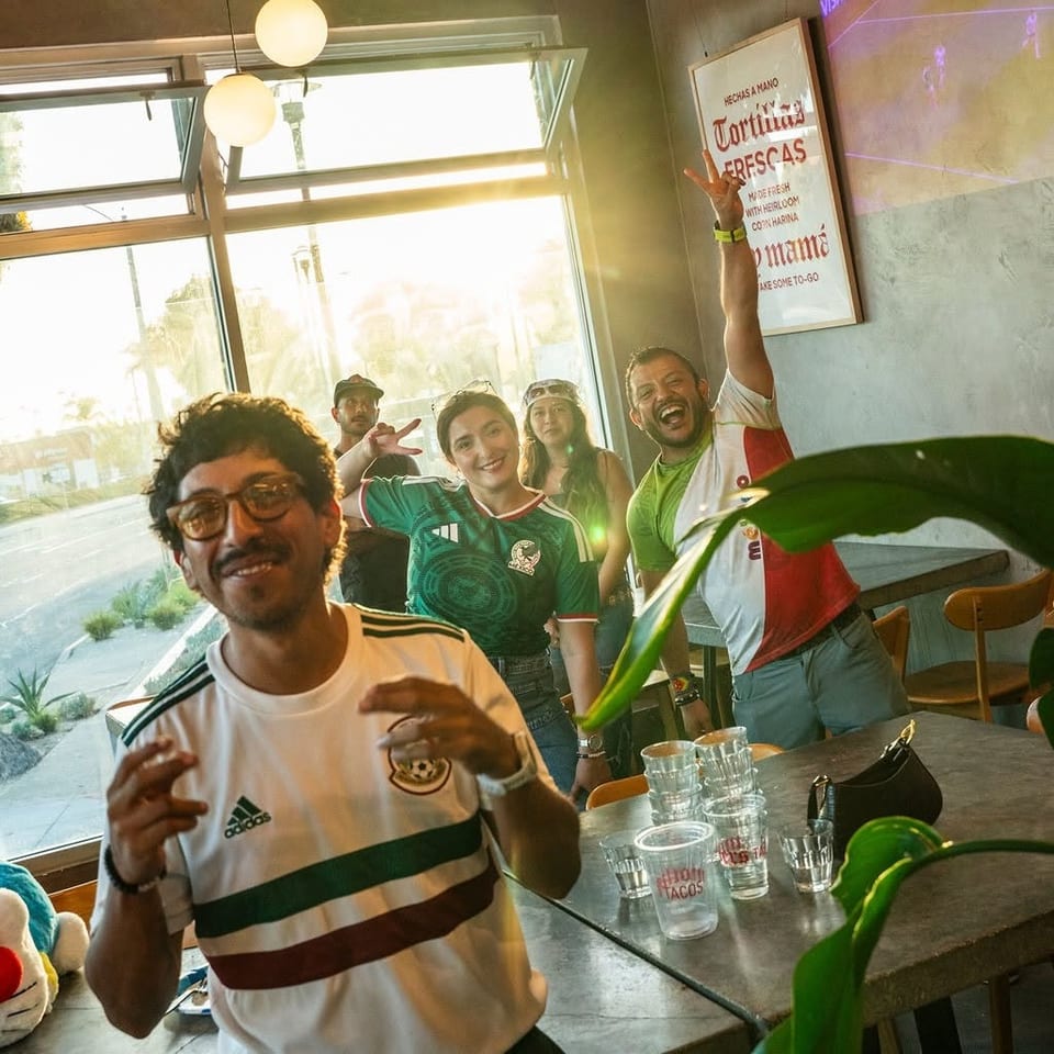 Friends in soccer jerseys celebrate inside a lively restaurant beside a table of drinks and a sign advertising fresh handmade tortillas.
