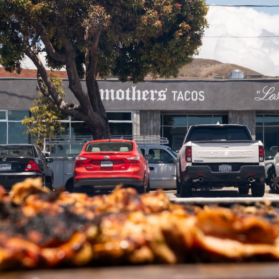 Mothers Tacos storefront with grilled food in the foreground.