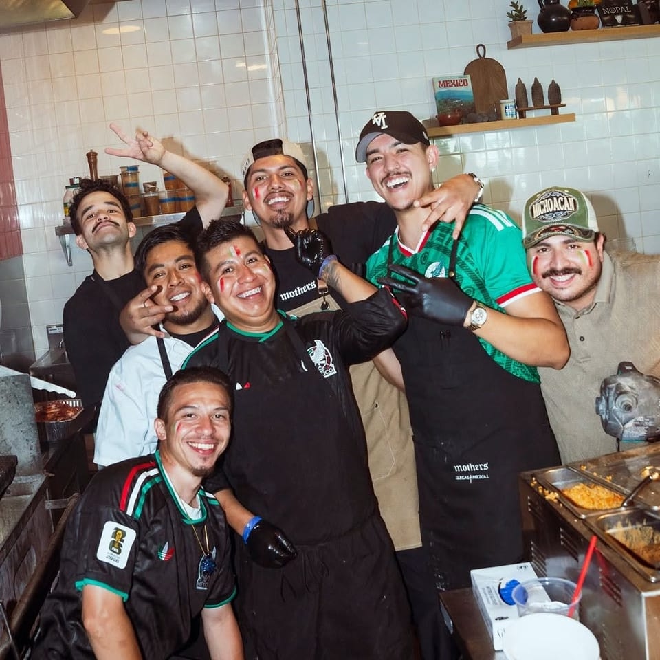 Smiling restaurant staff pose together in a tiled Mexican kitchen, wearing black aprons and colorful soccer jerseys beside food trays.