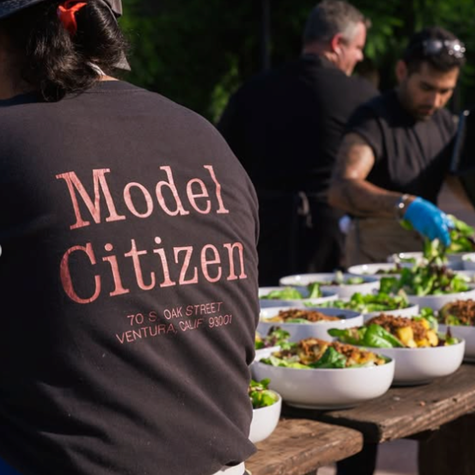 Volunteers prepare bowls of salad outdoors, while a person wearing a Model Citizen shirt stands in the foreground.