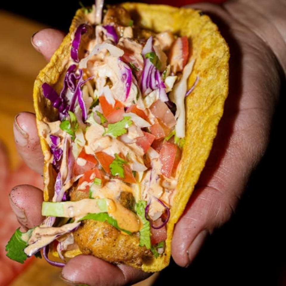 Handheld golden taco filled with seasoned meat, purple cabbage, lettuce, tomatoes, cilantro, and creamy sauce.