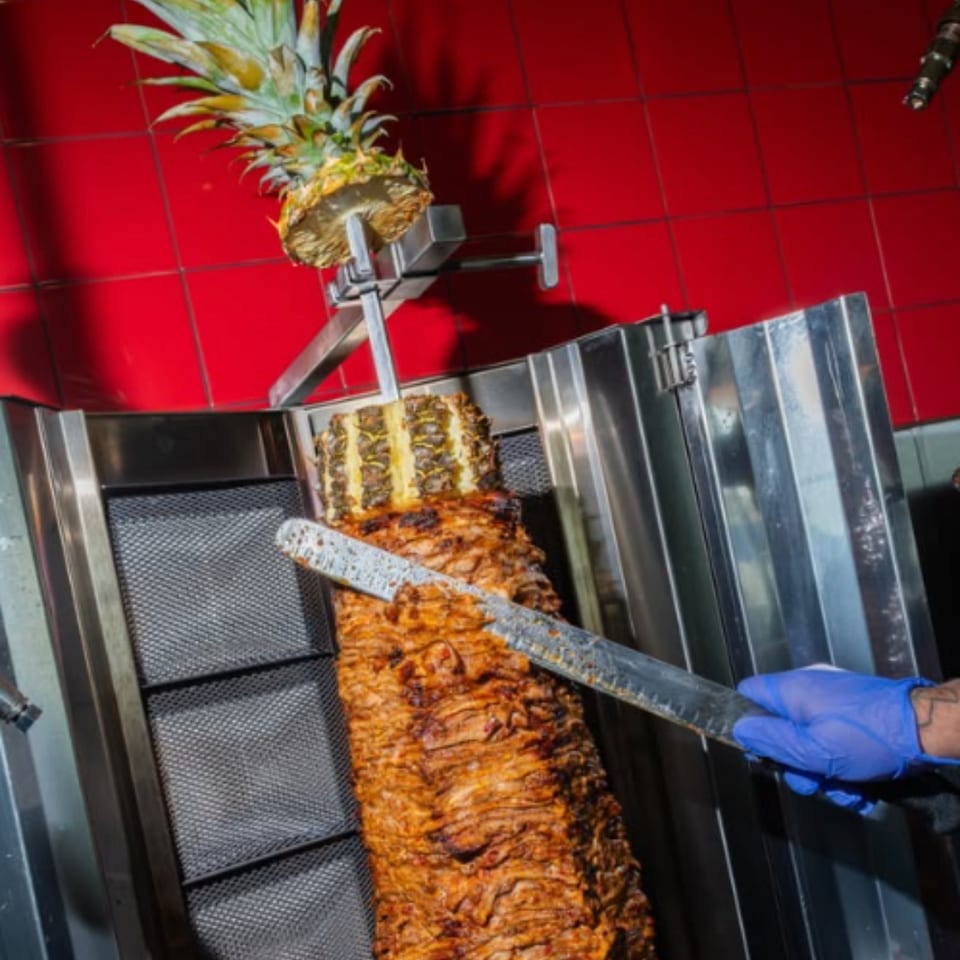Rotating spit of layered, grilled meat topped with a pineapple in a stainless-steel restaurant grill.